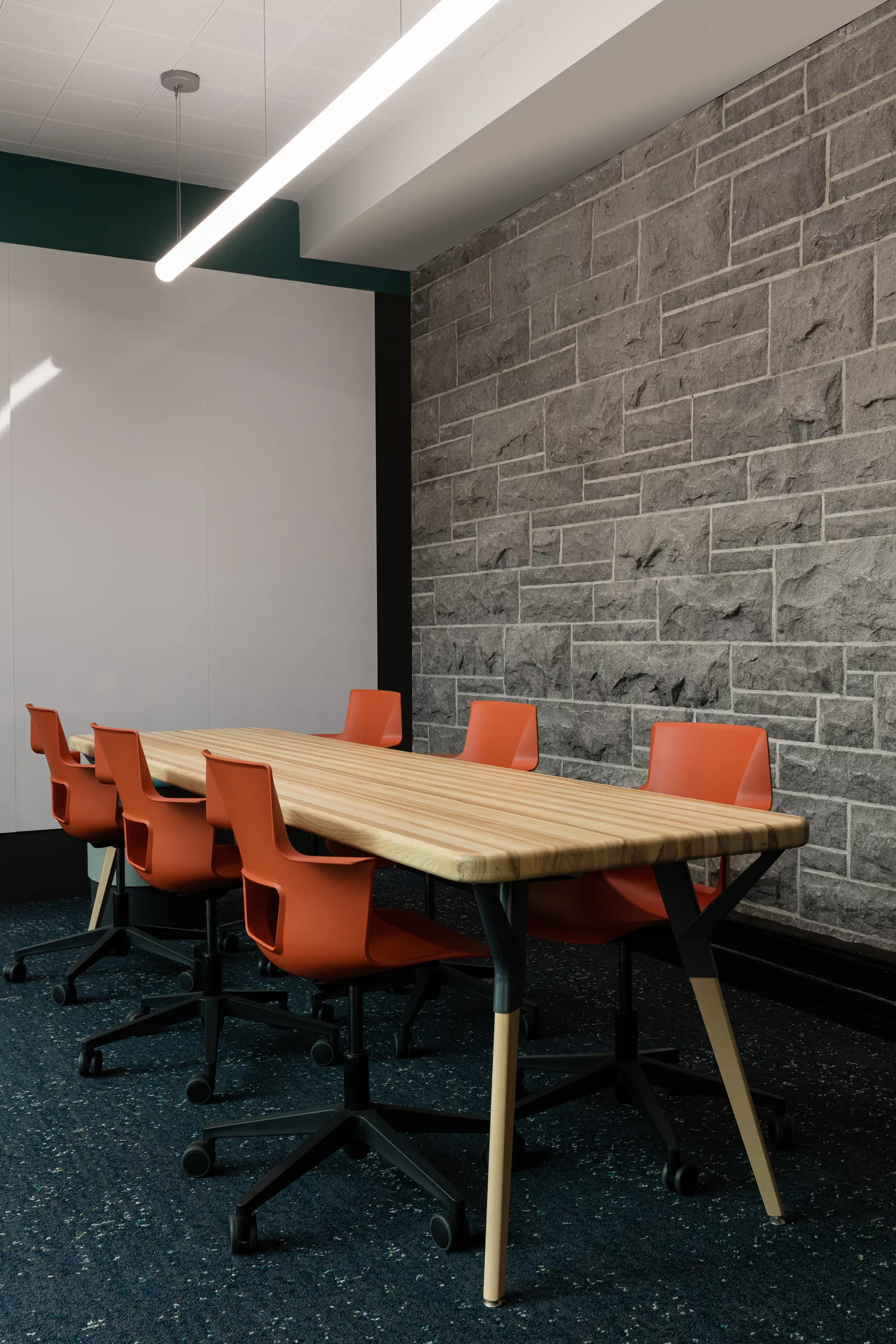 Une salle de réunion avec un mur de pierre au Collège-Notre-Dame avec une table en bois massif et des chaises de travail orange.