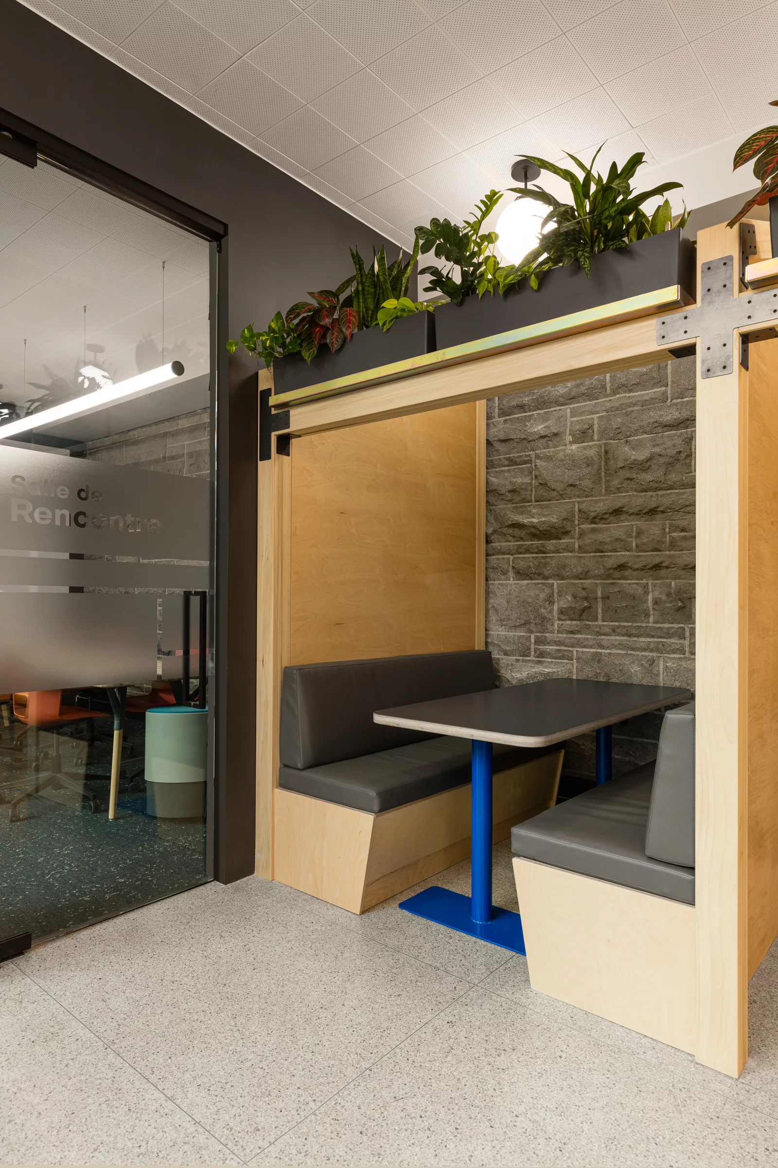 Une alcôve de travail avec une table et des banquettes intégrées, surplombée de plantes vertes. Les pieds de la table sont peints en bleu.