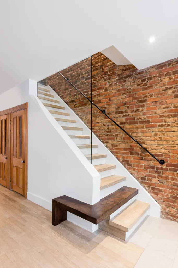 L'escalier qui monte au deuxième étage possède un mur vitré donnant sur la pièce du salon, aisi que la seconde marche qui est en bois plus foncé se transforme en petit banc sur le mur de gauche.