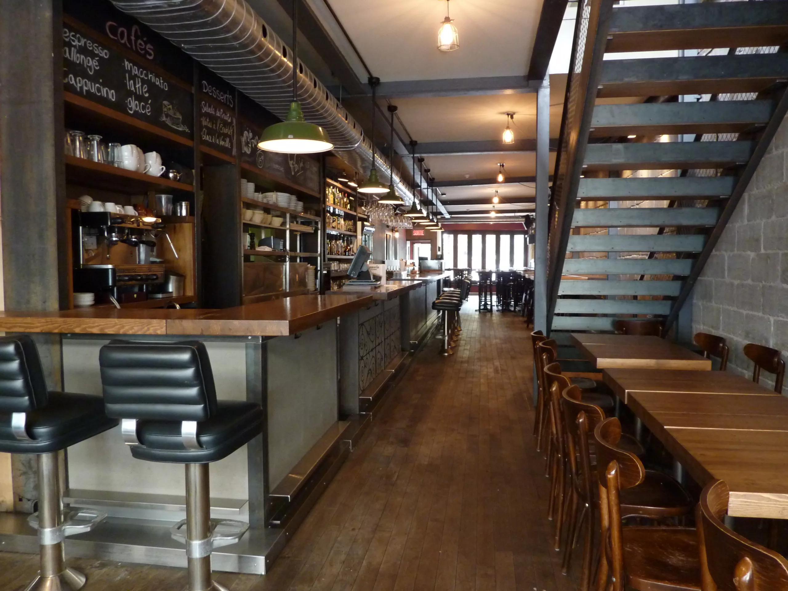 Vue de l'entrée du resto-bar Le Pourvoyeur, où l'on apreçoit des tables à droite sous l'escalier et le bar et ses tabourets du coté gauche.