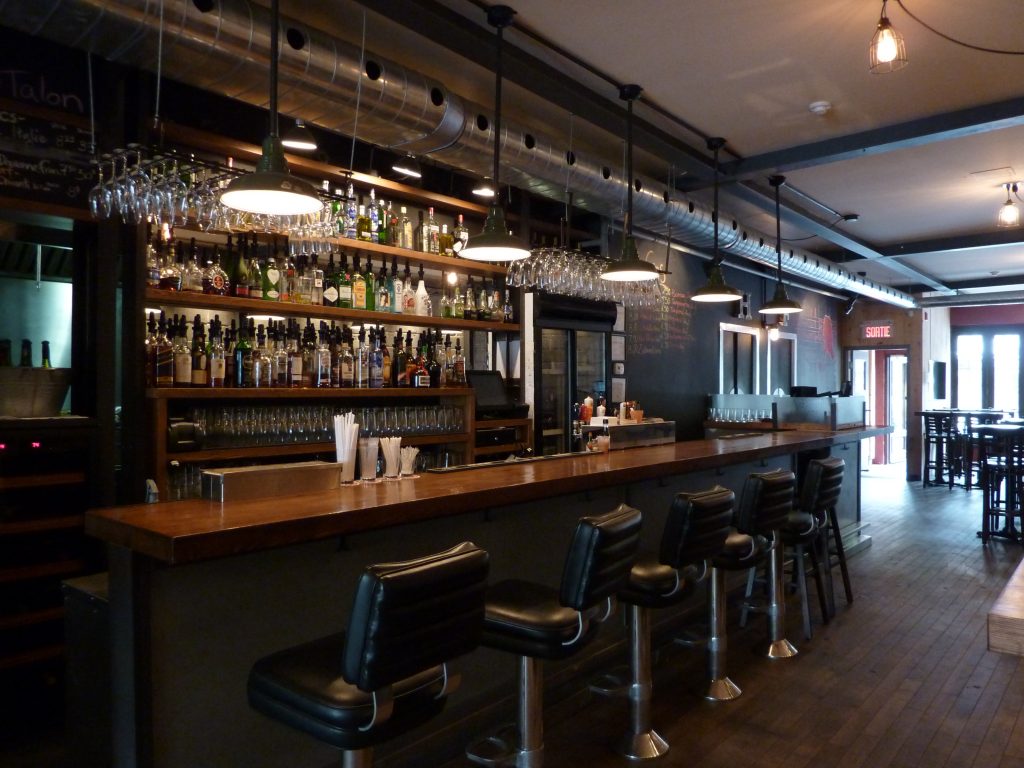 L'aménagement du bar du premier étage du Pourvoyer comprend un comptoir et des étagères de bois massif ainsi que des lampes industrielles suspendues. 