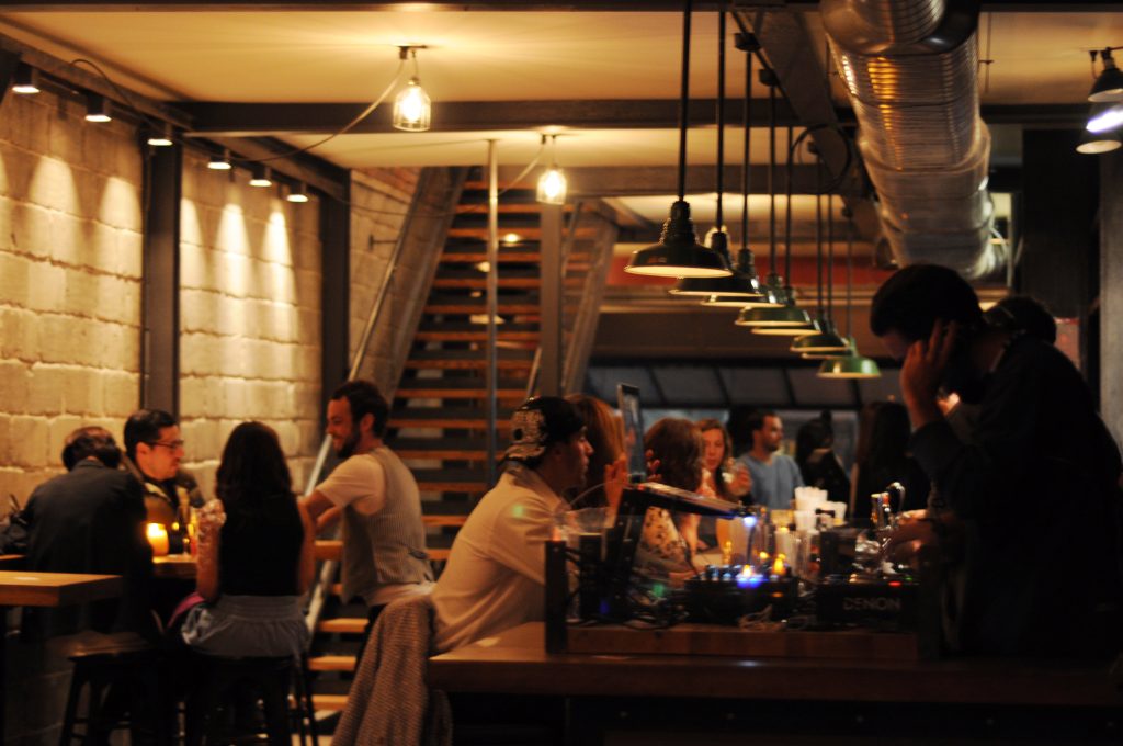 Le premier étage du resto-bar Le Pourvoyeur bien rempli lors d'une soirée festive, avec un DJ aux platines devant une rangée de lampes industrielles suspendues au dessus du bar.