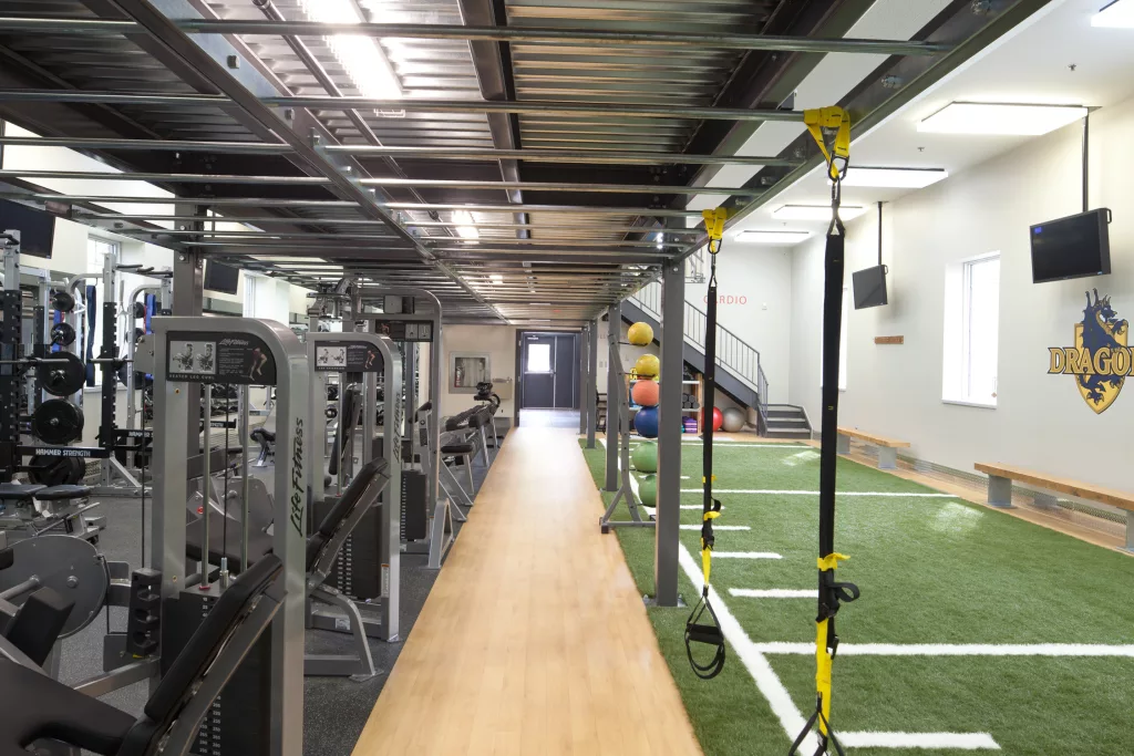 Une allé de bois sépare la section d'appareils de musculation de la salle d'entrainement d'une partie gazonnée d'un terrain de football.
