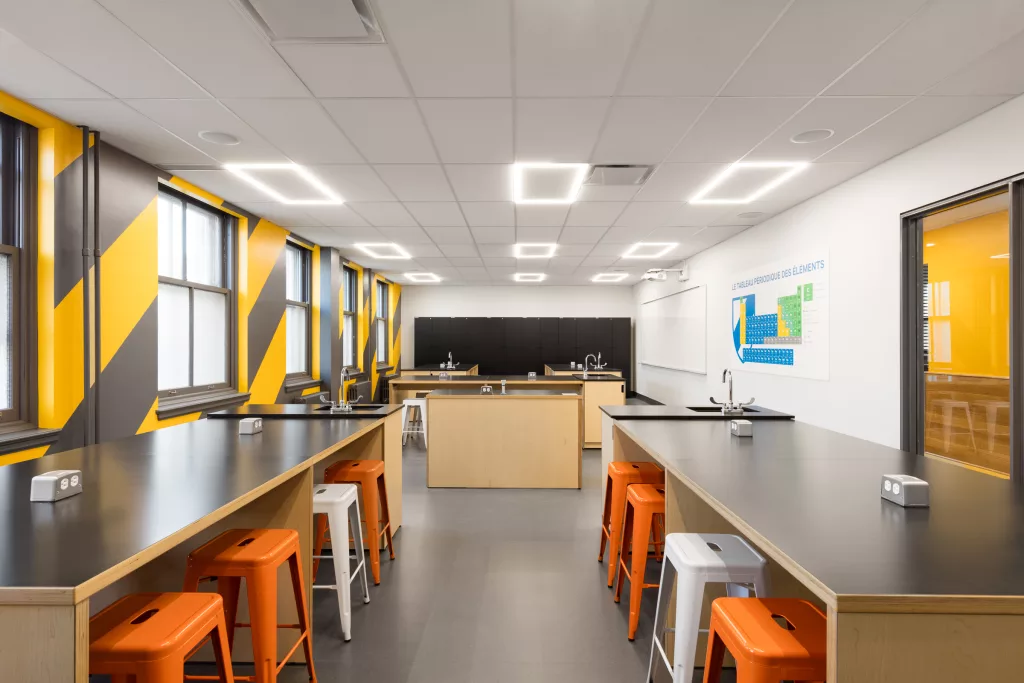 Le local de sciences physiques comprendd des tables de laboratoires de bois au dessus laminés noirs, les murs sont peints de diagonales jaunes et m=noires et les tabourets d'acier sont oranges et blancs.
