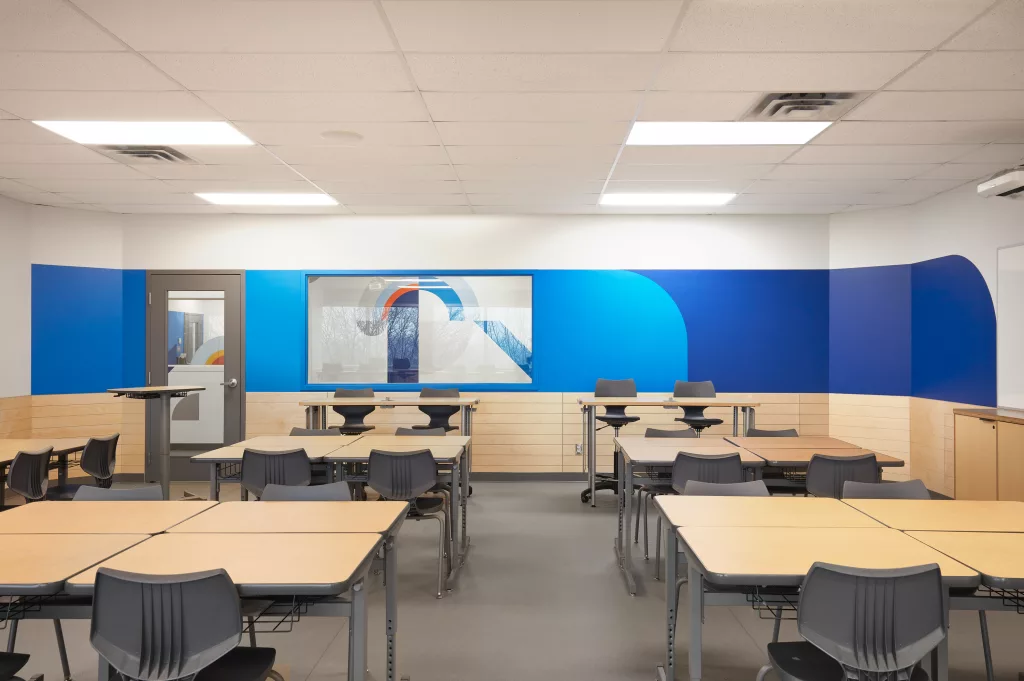 Une autre salle de classe de l'étage de deuxième secondaire consû par Taktk design comprends une fenetres lers le tiers lieu, une porte vitrée et des lattes de bois sur la partie basse des murs et du bleu sur le mur du fond, puis  des bureaux ajustables en hauteur et des chaises.