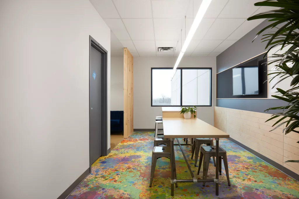 Un coin de l'étage au plancher coloré de taches de peinture comprend une table de bois sur deux niveaux, tes tabourets d'acier de tailles appropriées, un mur comprenant un tableau blanc et un téléviseur ainsi que des lattes de bois.