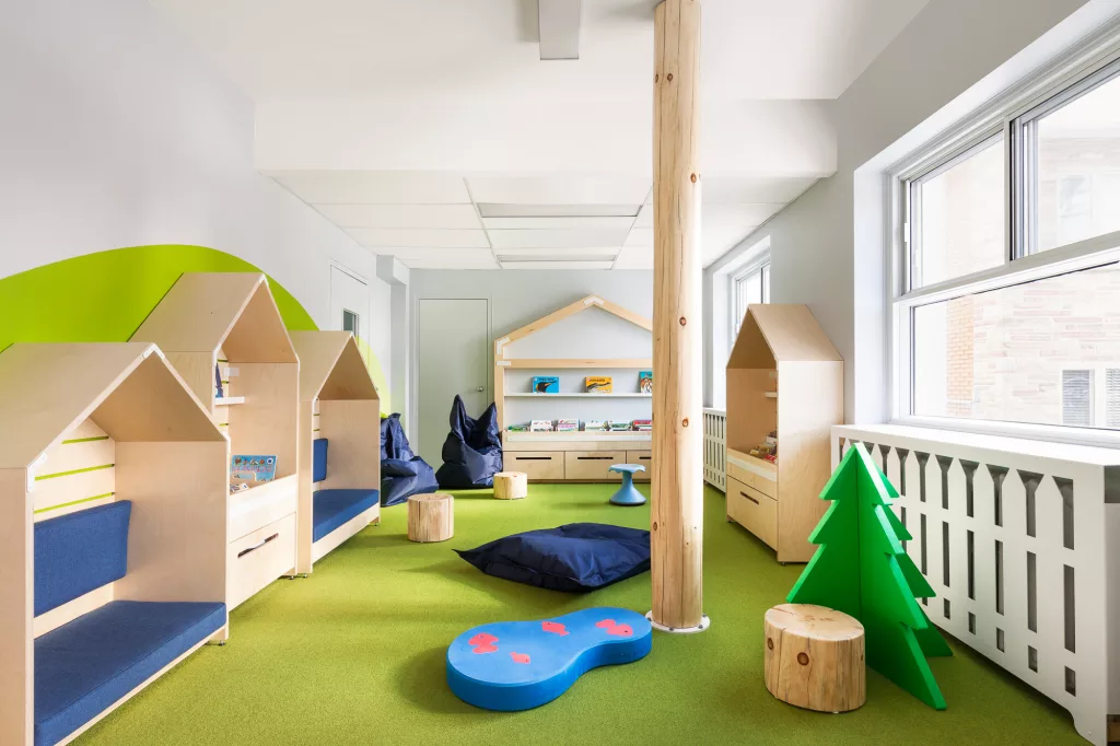Vue générale de la bibliothèque du Collège Ville Marie à Montréal. Un aménagement lumineux avec des couleurs naturelles et du bois clair. De petites cabanes en bois servent d'étagères pour les livres et de banquettes.
Un tronc d'arbre du sol au plafond donne une impression de nature à l'intérieur.