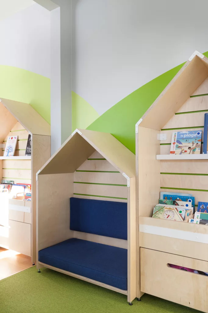 Vue rapprochée du mobilier de la bibliothèque. Un mobilier en forme de cabane créé un espace banquette cozy et intime pour la lecutre. Juste à côté une étagère reprenant la même forme crée une impression de petit village.