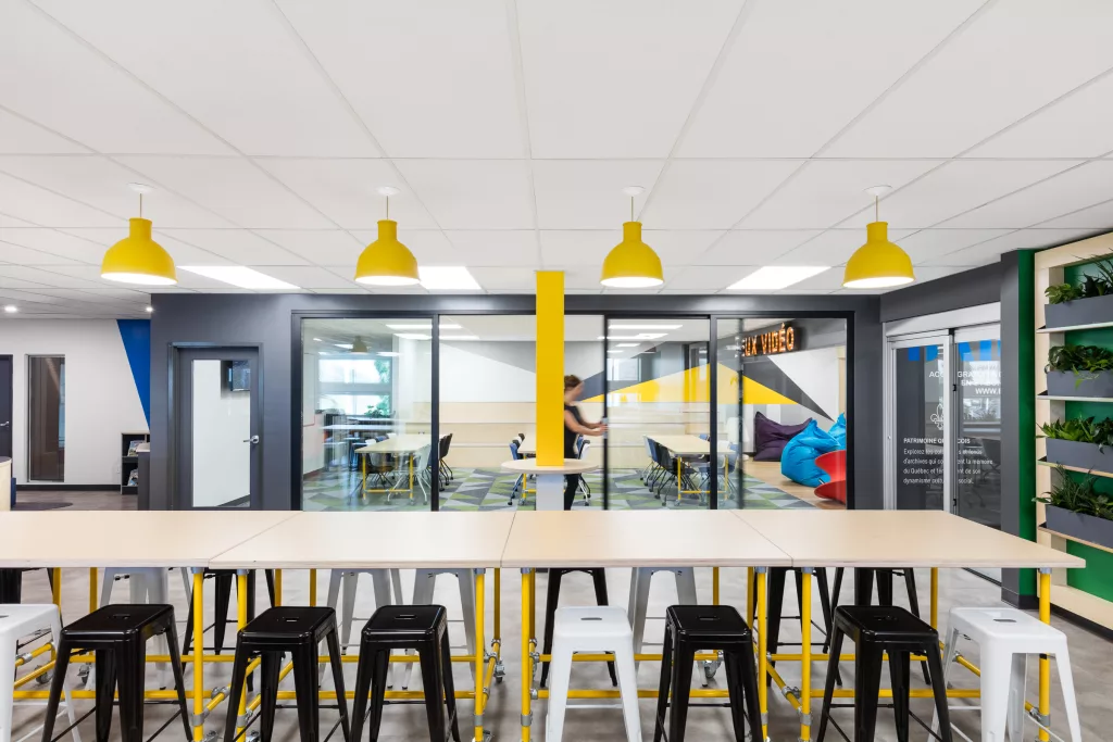 Au dessus des rangées de tables de bois sont suspendues des lampes industrielles de couleur jaune.