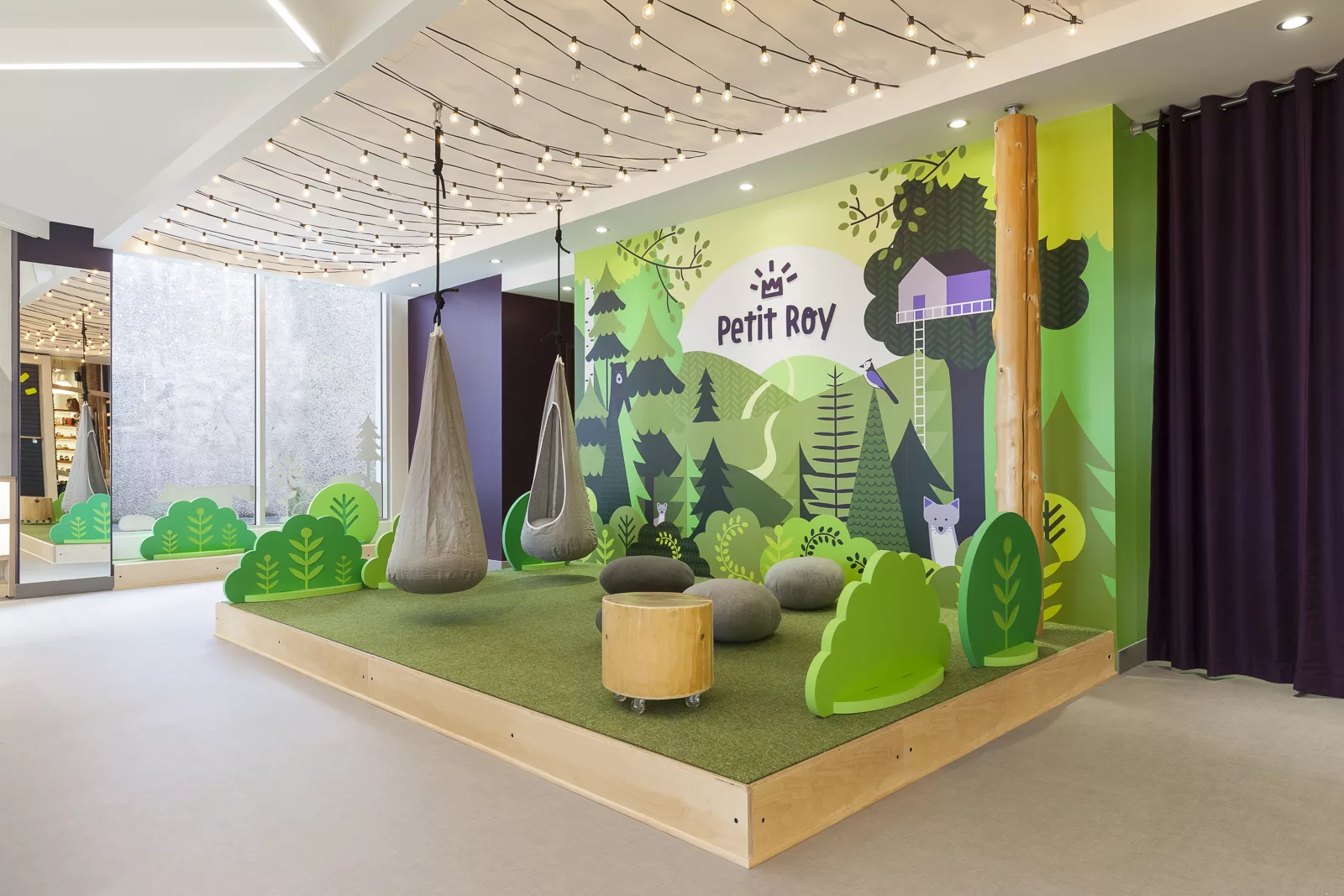 Vue de l'estrade de la boutique Petit Roy. Un aménagement évquant la nature, avec du bois, des éléments de décors vert et boisés, et une murale représentant un paysage verdoyant.