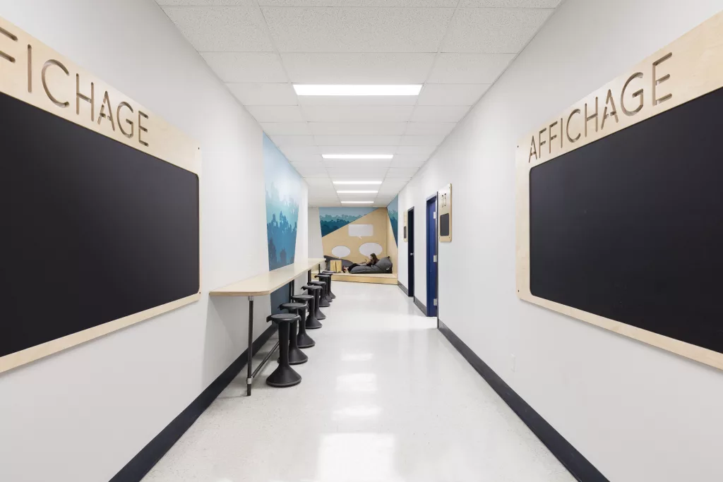 Un corridor meublé de tableaux d'affichages, d'un comptoir et de chaises tabourets et d'un coin de lecture avec des chaises poires.