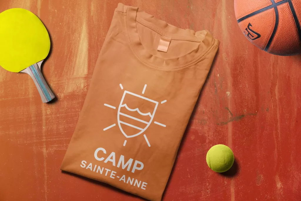 Un t-shirt orangé au logo blanc du camp Sainte-Anne entouré d'une raquette de pig-pong, d'un ballon de basketball et d'une balle de tennis.