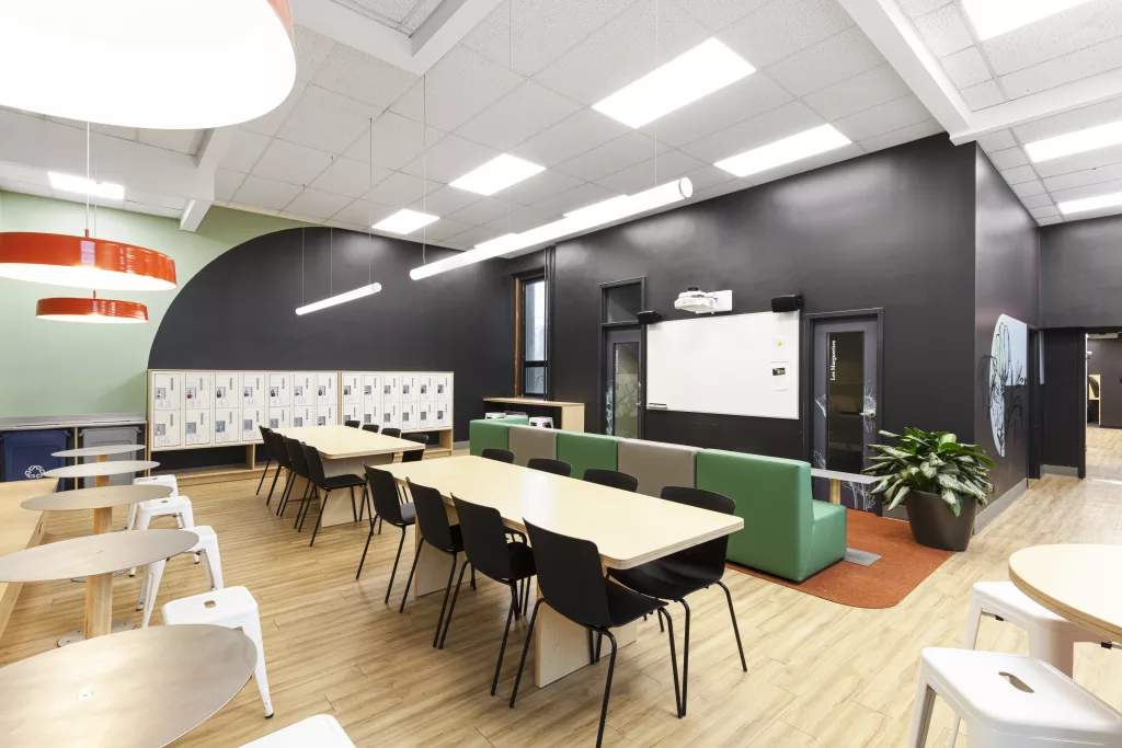 Cette classe flexible du Pensionnat du Saint-Nom-de-Marie comprend du mobilier de bois, des banquettes coussinées, un tableau blanc sur un mur noir et un projecteur.